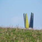 Die Spitze des Regionalpark-Turms ragt über einem Hügel hervor. Die Spitze besteht aus 4 Segmenten die wie zu einer Krone angeordnet sind. Der Hügel ist mit Graß bewachsen und der Himmel ist wolkenlos Blau.