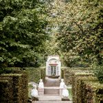 Weg im Schlossgarten Schwetzingen, er führt auf ein Gebäude zu von dem nur die Tür zu sehen ist, Auf beiden Seiten des Weges sind gerade geschnittene Hecken, auf halben Wege führt der Weg über eine Treppe, die Beidseitig von Sphinx-Skulpturen begrenzt wird.