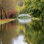 Ein Kanal, am Ufer stehen Bäume mit Herbstlaub, die sich im Wasser des Kanals spiegeln, Eine Weiße Bogenbrücke überspannt den Kanal, auch sie spiegelt sich im Wasser.
