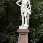Die Skulptur Geomethria im Schlossgarten Schwetzingen. Eine Weiße Marmorstatue steht auf einem Sockel aus rotem Sandstein. Die Figur ist nackt und hat nur ein Tuch locker um die lenden geschlungen, in der hand hält sie Messinstrumente.