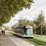 Eine Straßenbahn fährt auf den Gleisen die neben einem Füßweg verlaufen auf dem zwei Menschen gehen.