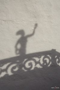 Schatten einer Figur und eines Geländers auf einer grob verputzten weißen Wand.