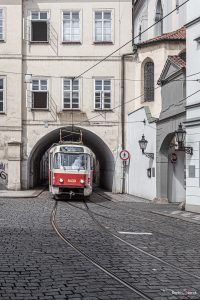 Eine rote Straßenbahn fährt durch ein Tunnel, der mit einem Haus überbaut ist, sie kommt auf den Betrachter zugefahren. Die Schienen sind in einer Straße aus Kopfsteinpflaster verlegt.