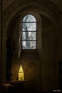 Hochkant Farbfoto. Durch ein Kirchenfenster fällt natürlichen Licht in einen dunklen Raum, als zweite Lichtquelle im Raum ist in der unteren linken Ecke eine Laterne in Form eies Hauses zu sehen, diese Lichtquelle erleuchtet ein Buch das direkt neben ihr liegt.