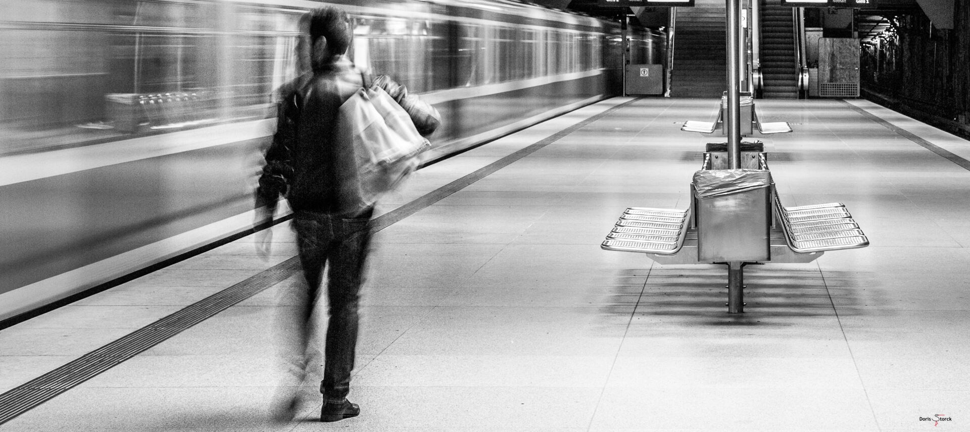 Schwarz-Weiß Foto von einem Bahnsteig eines U-Bahnhofs. Auf der linken Seite ist ein fahrender Zug zu erkennen, davor auf dem Bahnsteig läuft eine Person mit einer Tasche über der Schulter und Blick zum Zug, Richtung Ausgang. Zug und Person sind durch ihre Bewegung unscharf. Die Umgebung ist scharf. Auf dem Bahnsteig sind Bänke aus Metall die Rücken an Rücken zueinander stehen, Seitlich an jeder Bank befindet sich ein Mülleimer. Der Bahnsteig wird durch große runde Lampen an der Decke beleuchtet, im Hintergrund sind zwei Anzeigetafeln, auf denen sich die nächsten Verbindungen erahnen lassen. Am Ende des Bahnsteiges führen links eine Treppe und rechts eine Rolltreppe nach oben.