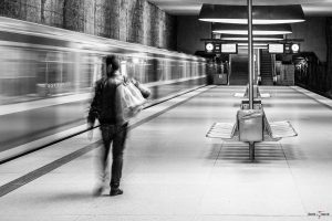 Schwarz-Weiß Foto von einem Bahnsteig eines U-Bahnhofs. Auf der linken Seite ist ein fahrender Zug zu erkennen, davor auf dem Bahnsteig läuft eine Person mit einer Tasche über der Schulter und Blick zum Zug, Richtung Ausgang. Zug und Person sind durch ihre Bewegung unscharf. Die Umgebung ist scharf. Auf dem Bahnsteig sind Bänke aus Metall die Rücken an Rücken zueinander stehen, Seitlich an jeder Bank befindet sich ein Mülleimer. Der Bahnsteig wird durch große runde Lampen an der Decke beleuchtet, im Hintergrund sind zwei Anzeigetafeln, auf denen sich die nächsten Verbindungen erahnen lassen. Am Ende des Bahnsteiges führen links eine Treppe und rechts eine Rolltreppe nach oben.