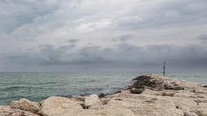 Farbfoto; ein Blick auf das Meer, auf der Wasseroberfläche kreuseln sich die Wellen. Die Horizontlinie läuft durch das untere Drittel, des Bildes, der Himmel ist trüb und die dunklen Wolken hängen tief. von Rechts ragt eine Buhne aus dicken Felsbrocken ins Bild. Am Ende der Buhne steht ein grünes Leuchtfeuer.