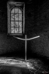 Schwarz Weiß Fotografie. In einem dunklen Raum, steht ein einfaches Holzkreuz. Von rechts fällt Licht auf das Kreuz. links über dem Kreuz ist in der Wand ein Rundbogenfenster, durch das äste eines Baums zu erahnen sind.