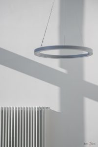 Hochkant Farbfoto, die dominierende Farbe ist weiß. In der oberen rechten Hälfte hängt eine reifenförmige silberne Lampe an drei dünnen Schnüren. Der Blick fällt von unten seitlich auf die Lampe. In der unteren linken Ecke ist ein weißer Heizkörper zu sehen. Die Wand im Hintergrund ist weiß gestrichen. Ein Kreuzförmiger Schatten fällt auf die Wand.